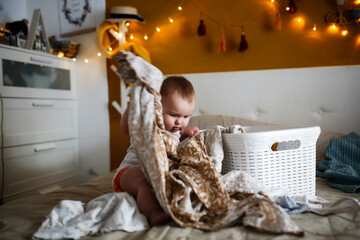 funny european chubby kid pulling out clothes from laundry basket on bed in cozy bedroom, mom help and home routines, motherhood and baby care, clean clothes and baby playing, childhood and emotions