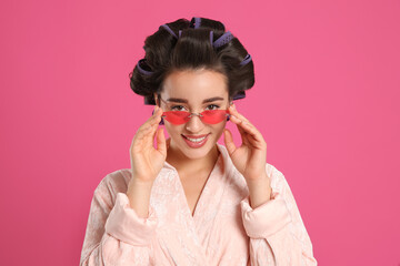 Happy young woman in bathrobe with hair curlers and sunglasses on pink background