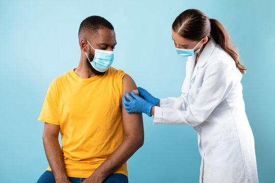 Doctor Putting On Band Aid On African American Male Patient After Coronavirus Vaccine Shot On Blue Studio Background