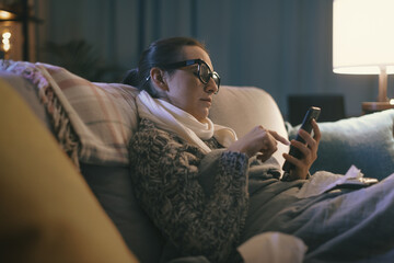 Obraz premium Sick woman resting on the couch and connecting with her smartphone
