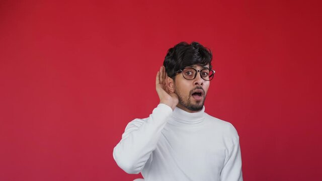 Hearing loss. Deafness disorder. Communication difficulty. Disturbed curious guy touching ear eavesdropping listening gossip isolated on red copy space background.