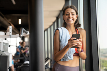 Fototapeta na wymiar Portrait of a cheerful sports woman using smartphone in fitness gym