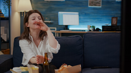 Person watching comedy on TV and eating delicious burger relaxing at home after work. Businesswoman having takeaway food on table while laughing, enjoying free time and takeout meal