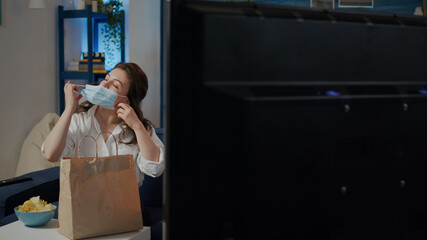 Woman with face mask sitting on couch with food delivery eating in living room at home while starting TV with remote. Young adult ordering takeaway meal package feeling hungry