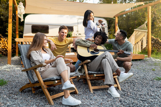 Young multiracial friends playing guitar and singing, holding dog, camping together, having fun autumn vacation outdoors