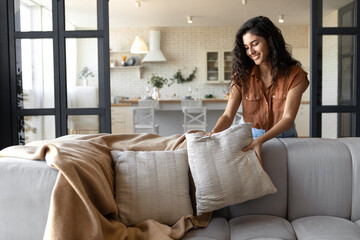 Lovely young woman putting soft pillows and plaid on comfy sofa, making her home cozy and warm,...