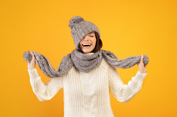 Overjoyed young lady in warm winter clothes pulling down knitted hat and playing with scarf on...