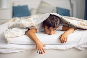boy child is lying in bed under striped blanket and does not want to sleep , insomnia and fun