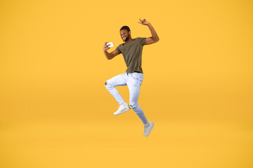 Fototapeta na wymiar Crazy african american man making selfie on smartphone while jumping in the air over yellow studio background