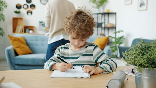 Slow motion of bored child doing homework writing sitting at desk while mother walking in house in background. Childhood and education concept.