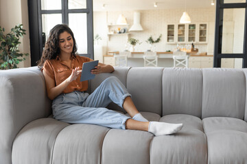 Millennial lady using tablet pc, studying or working remotely, having online conference, sitting on couch at home