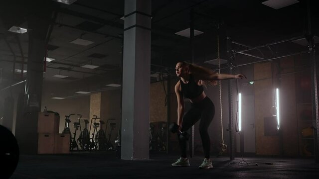A strong woman makes efforts and overcoming difficulties lifts a dumbbell in a dark gym. Fitness woman lifting weight dumbbells training in gym club.