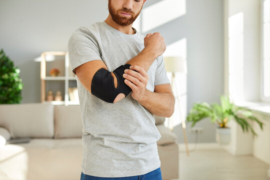 Man wears bandage on his elbow which serves as fixator to restore and relieve joint pain. Cropped image of man kneading his sick elbow and injured arm while standing at home. Pain relief concept.