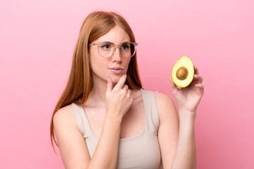 Young redhead woman holding an avocado isolated on pink background having doubts