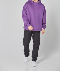 Cropped shot of man in purple blank hoodie and black pants. Standing on gray background. Mockup for...