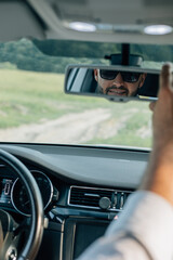 Bearded caucasian business man looking at camera through rear view mirror. Head and shoulders. Summer background.