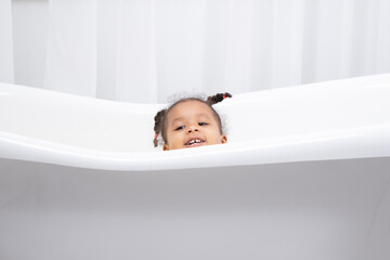 A funny little African girl looks out from a white bathtub and smiles.