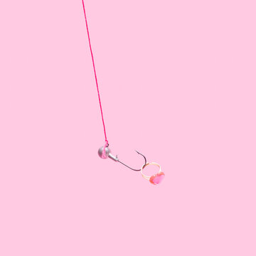 Minimal concept of catching love. Arrangement made of a shiny ring with a heart shaped pink gem on a fish hook. Soft pop pink background.