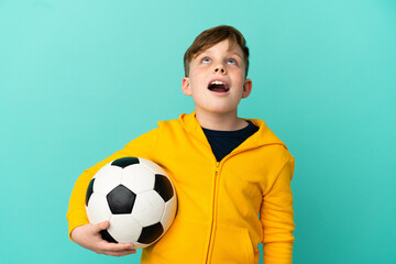 Redhead kid playing football isolated on blue background looking up and with surprised expression