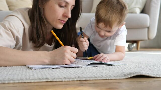 A little boy and a loving mother are sitting on a warm floor in a cozy living room, holding colored pencils and coloring pictures on a paper sheet.Child development concept