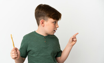 Little redhead boy holding a toothbrush isolated on white background surprised and pointing finger to the side