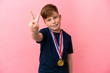 Little redhead boy with medals isolated on pink background smiling and showing victory sign