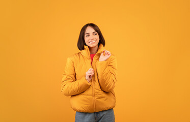 Cheerful young woman wearing and zipping up warm jacket, smiling and looking aside at free space, orange background © Prostock-studio