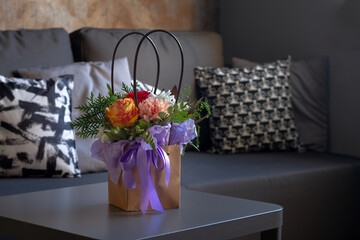 A bouquet of flowers in a paper bag with a purple ribbon stands on a gray table in a room near a dark sofa