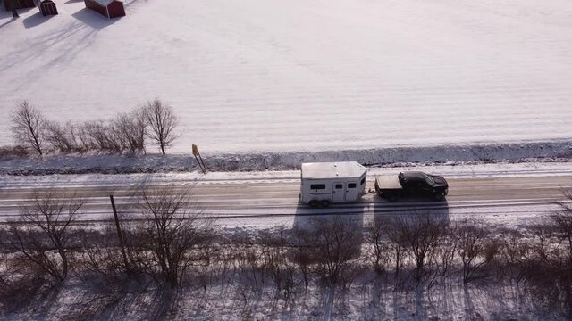 Truck towing white horse trailer on rural road in winter season, aerial drone shot