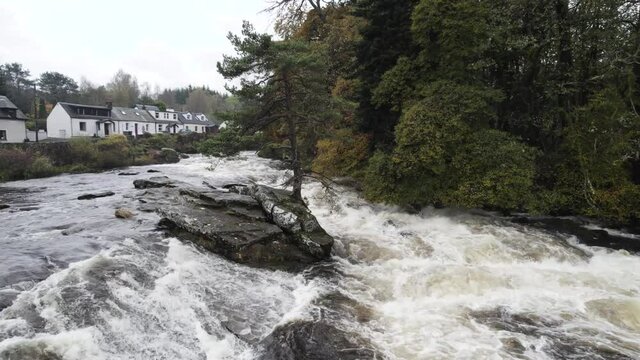 Aerial drone footage rolling slowly over fast flowing water and river rapids at the Falls of Dochart in Killin, Trossachs National Park, Scotland with white water, rocks and surrounded by native trees