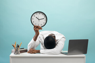 Tired african american businessman sitting at desk with pc laptop, holding clock, feeling...