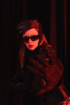 frowning woman in black sunglasses, beret and leather jacket showing refuse gesture on dark background.