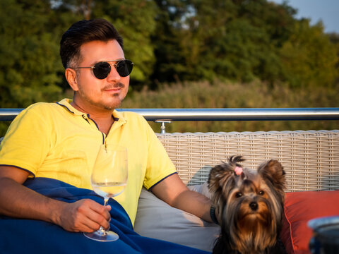 young black hair person wearing casual clothes and sunglasses sitting on a terrace wrapped in blanket with his yorkshire terrier dog and holding glass of wine, enjoying in a sunny summer evening