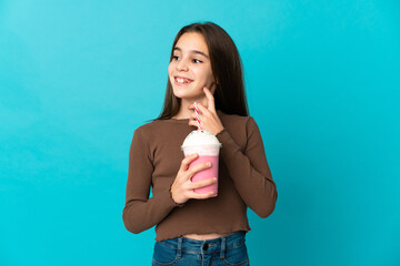 Little girl with strawberry milkshake isolated on blue background thinking an idea while looking up