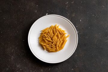 Raw penne macaroni on white plate on dark background. Cooking pasta concept.Top view