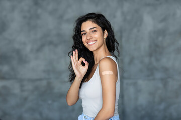 Covid vaccination. Young woman with band aid after coronavirus vaccine injection gesturing okay over grey background