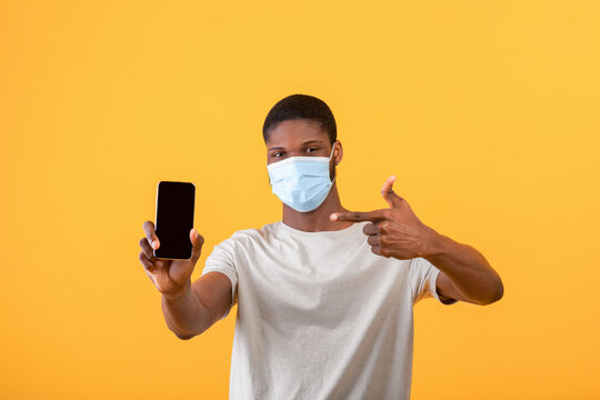 African american guy in protective medical mask on face pointing at smartphone with blank screen, yellow background - Powered by Adobe