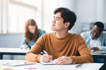 Smiling asian student sitting at desk in classroom thinking
