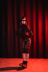 sexy woman in sunglasses, lace leggings, leather jacket and boots posing with hands on waist on dark draped background with red light.