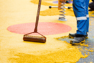 Worker leveling and pressing rubber coating for playgrounds with roller, mason hand spreading soft rubber crumbs. Outdoor soft coating and floor covering for sports. Selective focus. - obrazy, fototapety, plakaty