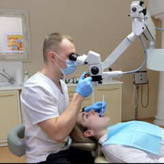 Naklejka na ściany i meble A young girl on a dental chair and a dentist who sits next to him. He looks at the teeth with a dental microscope and holds a dental bur and a mirror in his hands.