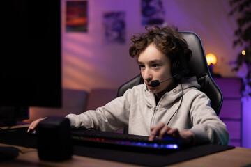 School-age boy pro gamer spends time playing computer games, focused child has headset to talk to team, room lit with neon, led lights, gaming accessories