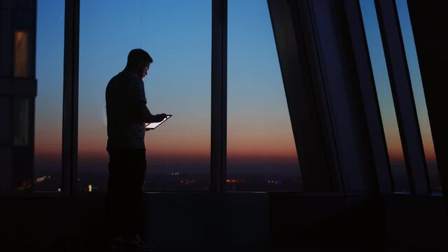 Silhouette of young man standing near window with tablet computer and looks at the sunset city through the windows of a skyscraper. Alone man in office at the end of the day