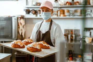 Employee in mask holds tray with fresh decorated croissants in bakery shop