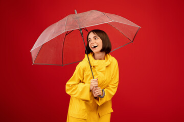 Stylish accessories. Playful woman standing under transparent umbrella, smiling and looking aside over red background