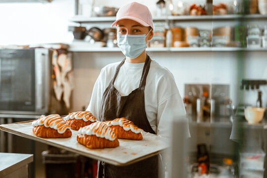 Employee in mask holds tray with fresh decorated croissants in bakery shop