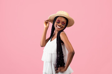 Stylish young black lady in lovely dress, sunglasses and straw hat posing and smiling on pink studio background