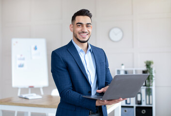Cheerful Arab businessman in elegant suit holding laptop pc, smiling at camera in modern office