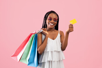 Easy shopping. Joyful Afro lady holding credit card and bright shopper bags on pink studio background