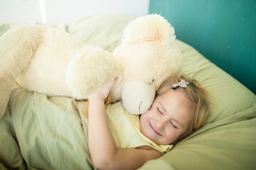 A little caucasian girl plays with a teddy bear in bed, hugs him and puts him to sleep